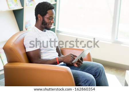 Young male student using digital tablet while sitting on armchair in bookstore