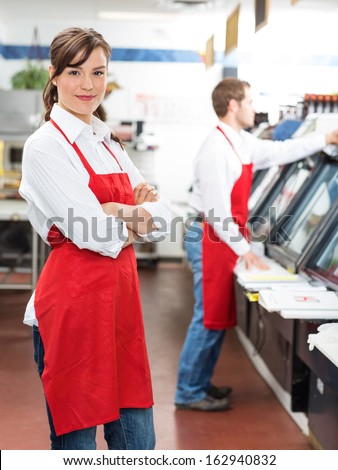 Portrait of female butcher standing arms crossed with colleague working in background at store