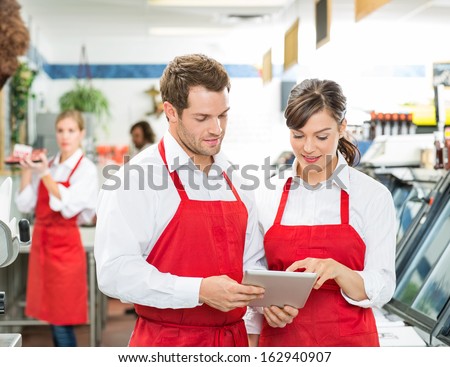 Male And Female Butchers Using Digital Tablet At Store With Colleagues Working In Background