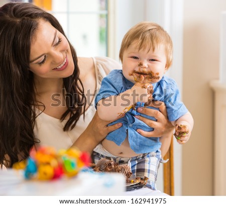 Mid adult mother holding baby boy eating cake with icing on face at birthday party