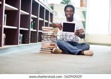Full length portrait of smiling young student with stacked books showing digital tablet while sitting on floor at library