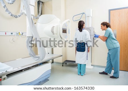 Full length of nurse adjusting xray machine for female patient in examination room
