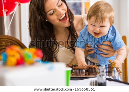 Cheerful mother holding baby boy with messy hands covered with cake icing at birthday party