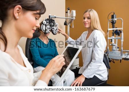 Female eye doctor examining senior woman\'s eyes while colleague using digital tablet in store