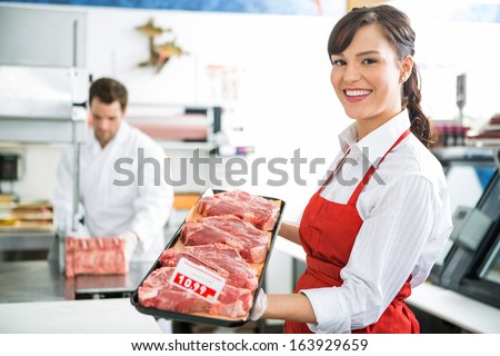 Portrait of happy female butcher holding meat tray in store with colleague working in background
