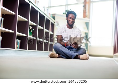 Full length of young male student using digital tablet while sitting on floor at library