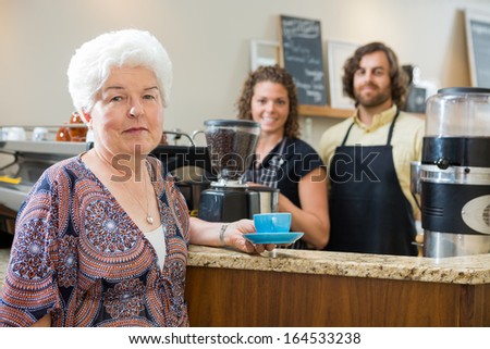Portrait of senior woman holding coffee cup with workers in background at cafe counter