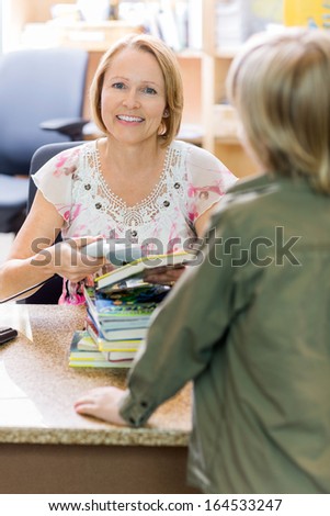 Portrait of happy mature librarian scanning books while boy standing at checkout counter in library