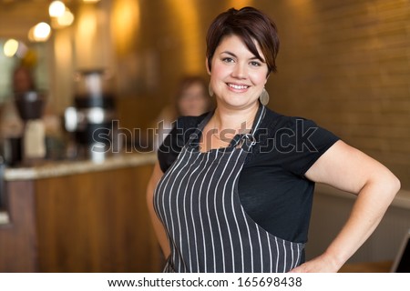 Portrait Of Happy Young Female Owner With Hands On Hips Standing In Coffee Shop