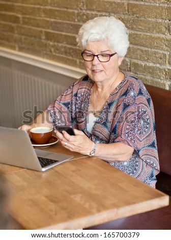 Senior woman with laptop and coffee cup text messaging through smartphone in cafe
