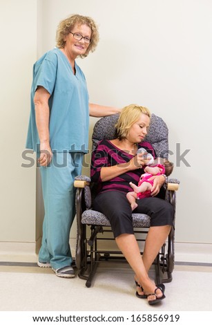 Full length portrait of nurse standing by mother feeding newborn babygirl in hospital