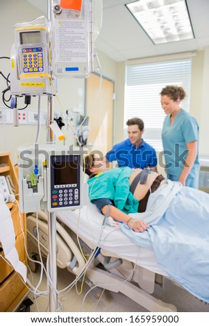 IV and epidural equipment with nurse and man looking at pregnant woman lying on bed in hospital