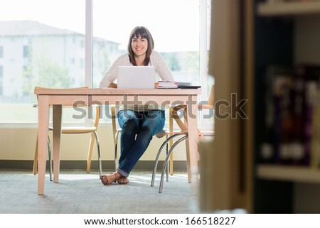 Full length portrait of smiling university student with laptop sitting at table in library