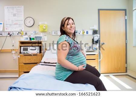 Side view portrait of happy pregnant woman with hands on stomach sitting on hospital bed