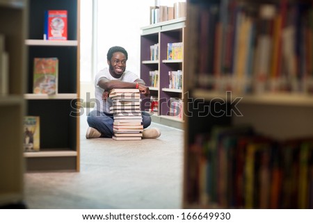 Full length of happy male student with stacked books looking away while sitting on floor at library
