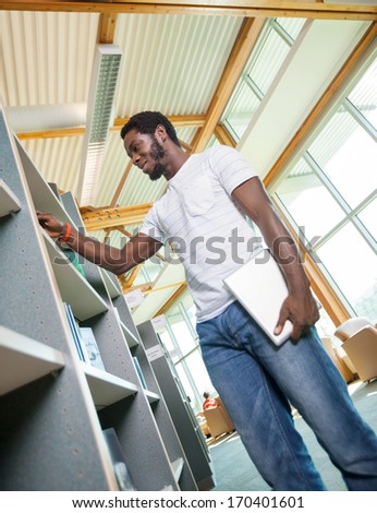 Low angle view of male student with digital tablet choosing book in bookstore
