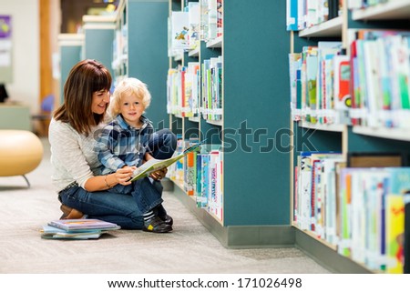 Portrait Of Boy With Teacher Reading Book By Bookshelf In Library