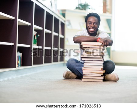 Full length portrait of happy African American student leaning on stacked books while sitting on floor at library