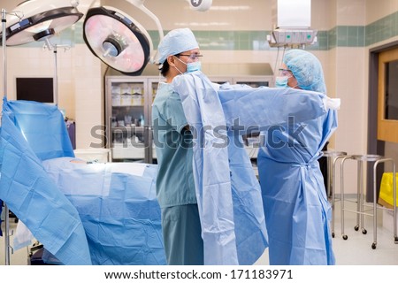 Side View Of Mature Scrub Nurse Assisting Surgeon Putting On Sterile Operating Gown