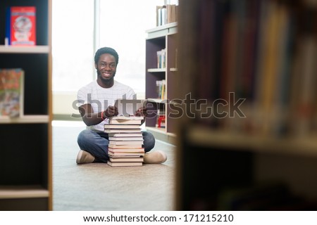 Full length portrait of male student with books and digital tablet sitting on floor at library