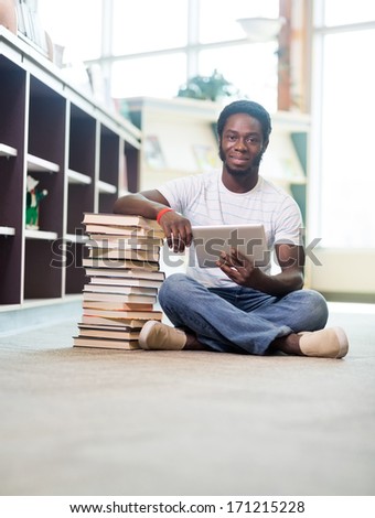 Full length portrait of African American male student with digital tablet and stacked books sitting on floor at library