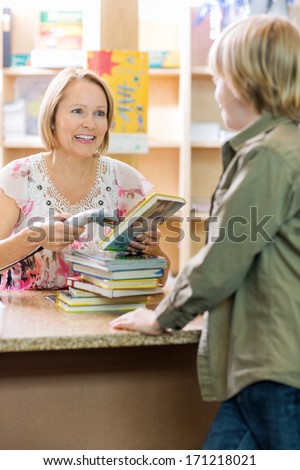 Young boy standing at checkout counter while librarian scanning books in library