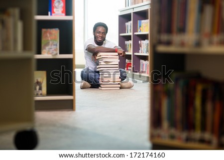 Full length portrait of male student with books and digital tablet sitting on floor at library