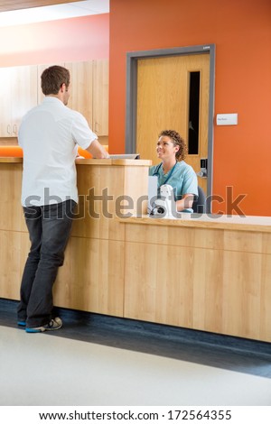 Full length of male patient conversing with nurse at reception desk in hospital