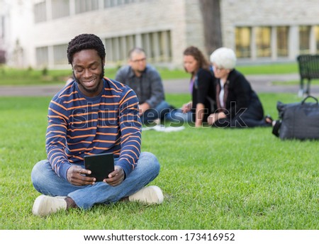 Male student using digital tablet on grass at campus park with friends studying in background