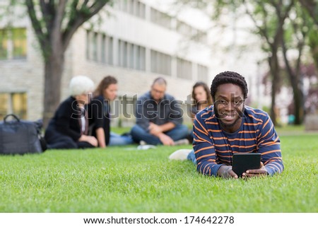 Portrait of student holding digital tablet on grass at campus park with friends studying in background