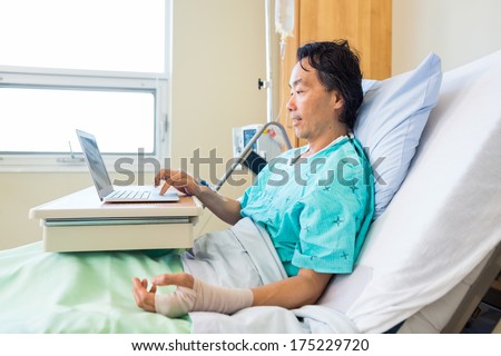 Side view of mature male patient with crepe bandage on hand using laptop on bed in hospital