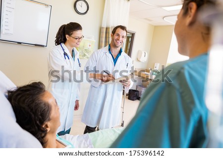 Happy young doctors discussing notes with patient and nurse in foreground at hospital room