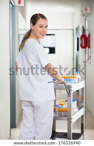 Portrait of happy young female lab technician pushing medical cart in corridor