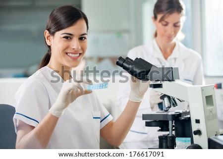 Portrait of young female researcher holding slide while using microscope in laboratory Portrait of young female researcher holding slide while using microscope in laboratory