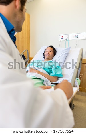 Smiling male patient looking at doctor writing on clipboard while lying on bed in hospital room