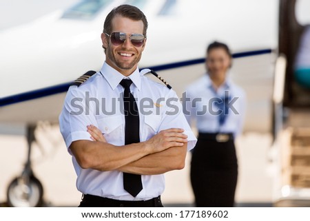 Portrait of confident pilot with arms crossed against stewardess and private jet at terminal