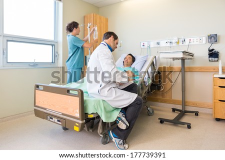 Doctor writing on clipboard while nurse adjusting IV drip on rod by patient\'s bed in hospital