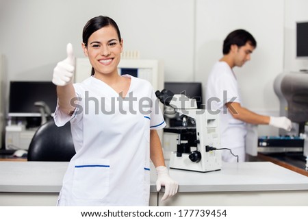 Portrait of happy young female scientist gesturing thumbs up with colleague in background at laboratory