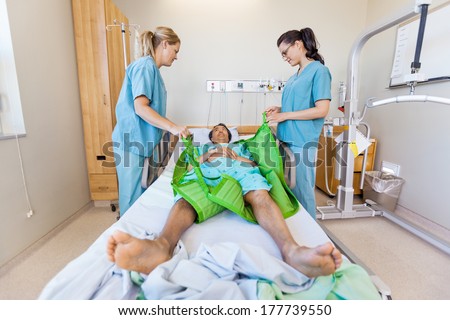 Nurses preparing male patient before transferring him on hydraulic lift on bed at hospital