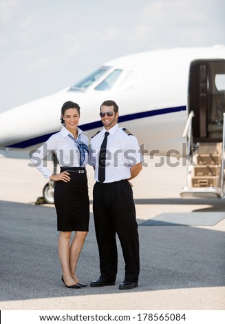 Full length portrait of confident stewardess and pilot standing against private jet at terminal