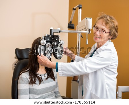 Portrait of senior female optometrist adjusting phoropter for young patient in eye clinic