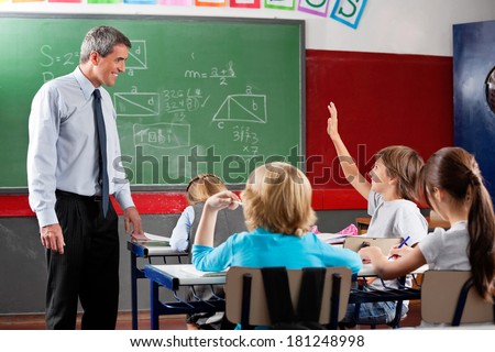 Happy mature professor looking at little schoolboy raising hand in classroom