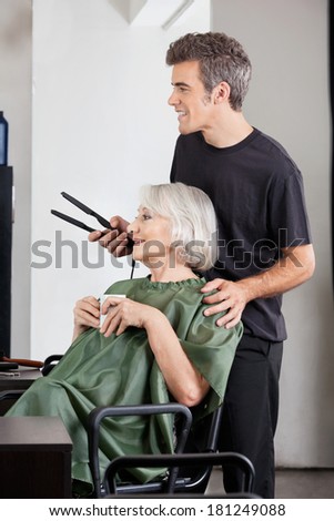 Senior woman with coffee cup and hairdresser holding straightener at beauty parlor