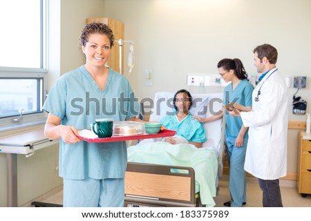 Portrait of confident nurse holding breakfast tray with medical team and patient in background at hospital