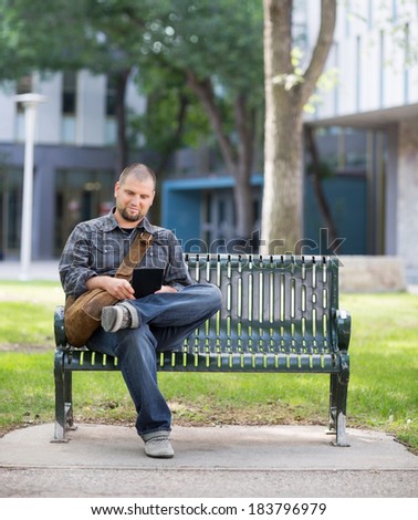 Mid adult male university student using digital tablet while sitting on bench at campus