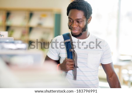 Smiling young male student carrying backpack while looking at books in bookstore