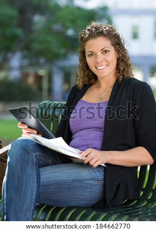 Portrait of happy female student with book and digital tablet sitting on bench at university campus