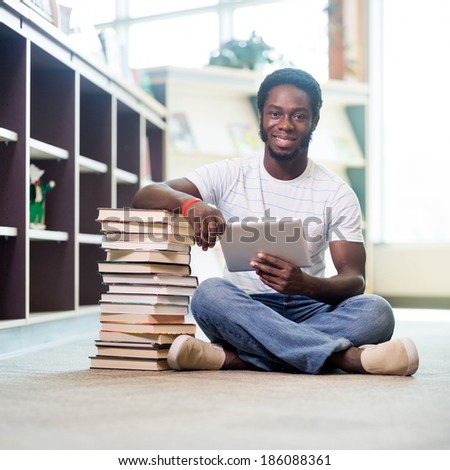 Full length portrait of young male student with stacked books and digital tablet sitting on floor at library