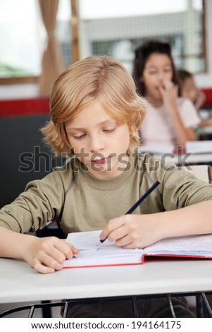 Cute little schoolboy writing in book at desk with classmates in background