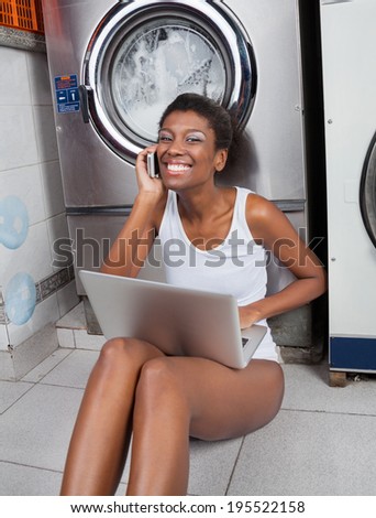 Portrait of happy young African American woman using laptop and mobilephone in laundromat